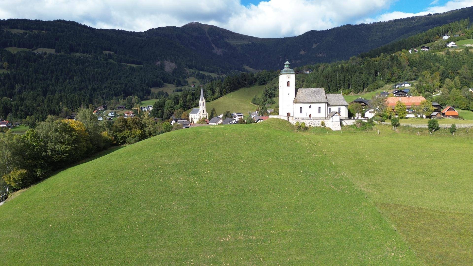 Panoramablick auf ein malerisches Dorf mit zwei Kirchen, eingebettet in eine grüne Hügellandschaft.