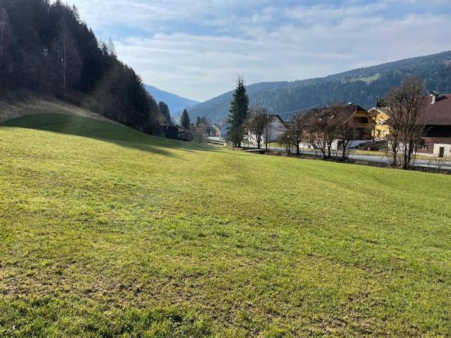 Sonnige, weitläufige grüne Wiese mit Blick auf Häuser und Berge unter blauem Himmel.