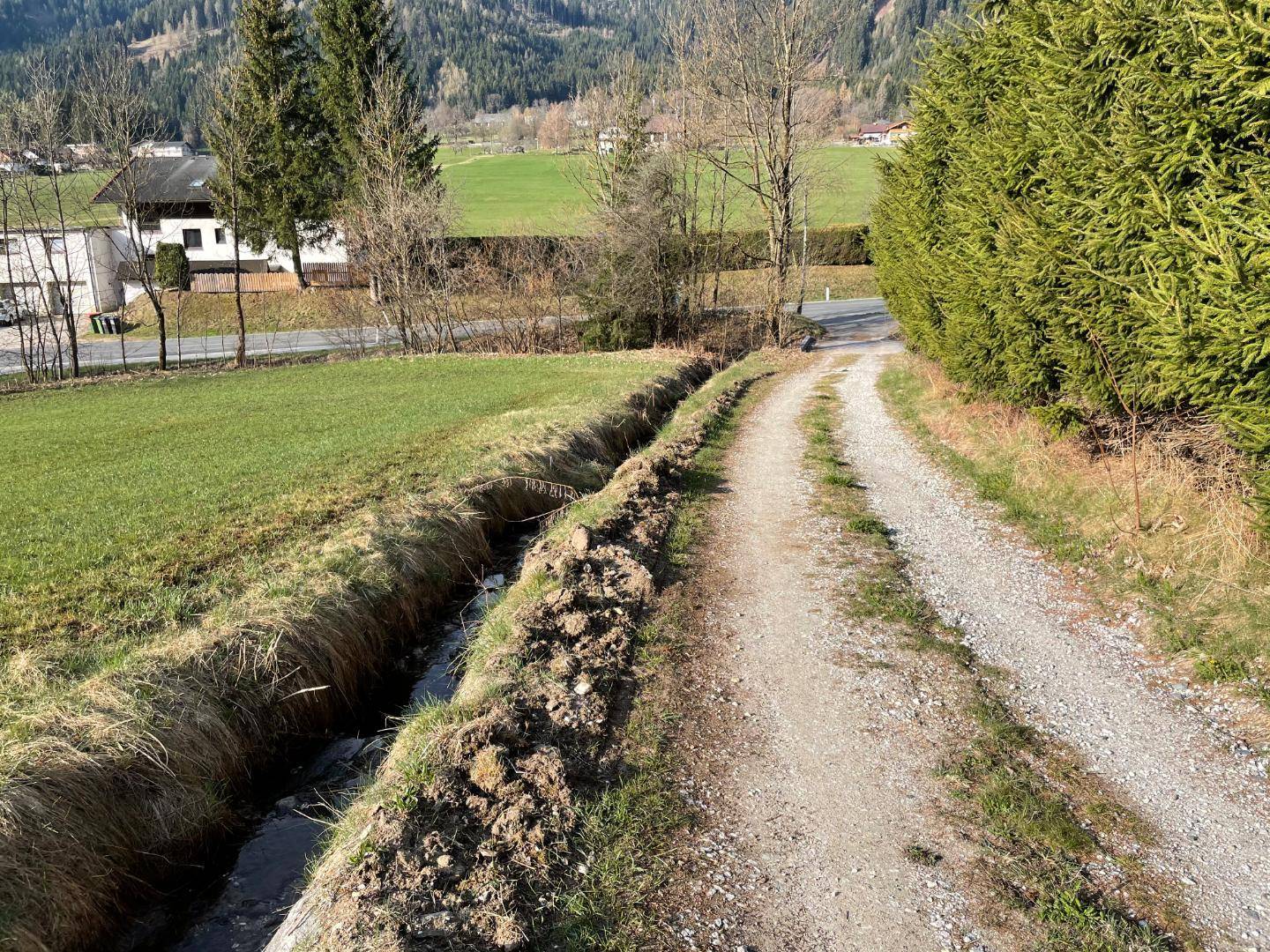 Schotterweg entlang eines kleinen Grabens mit grünen Wiesen, einer Hecke und Bergen im Hintergrund.