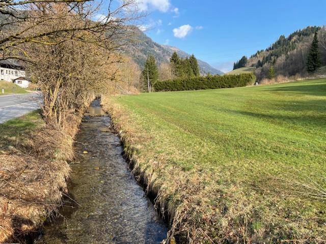 Kleiner Bach fließt neben einer Straße und einer weiten grünen Wiese mit Bergen im sonnigen Hintergrund.