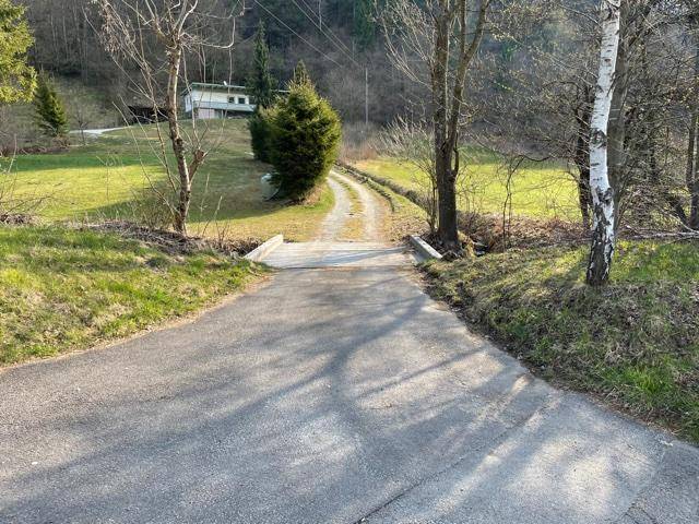 Asphaltierte Zufahrtsstraße führt über eine kleine Brücke zu einem Schotterweg und einem Haus am Hang.