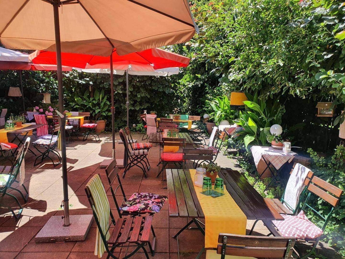 Weitläufige Terrasse mit zahlreichen Tischen, Stühlen und großen Sonnenschirmen unter grünen Bäumen an einem sonnigen Tag.