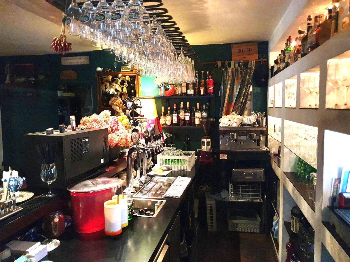 Voll ausgestattete Bar mit zahlreichen Flaschen, hängenden Gläsern und professioneller Ausstattung, vor dunkelgrünen Wänden.