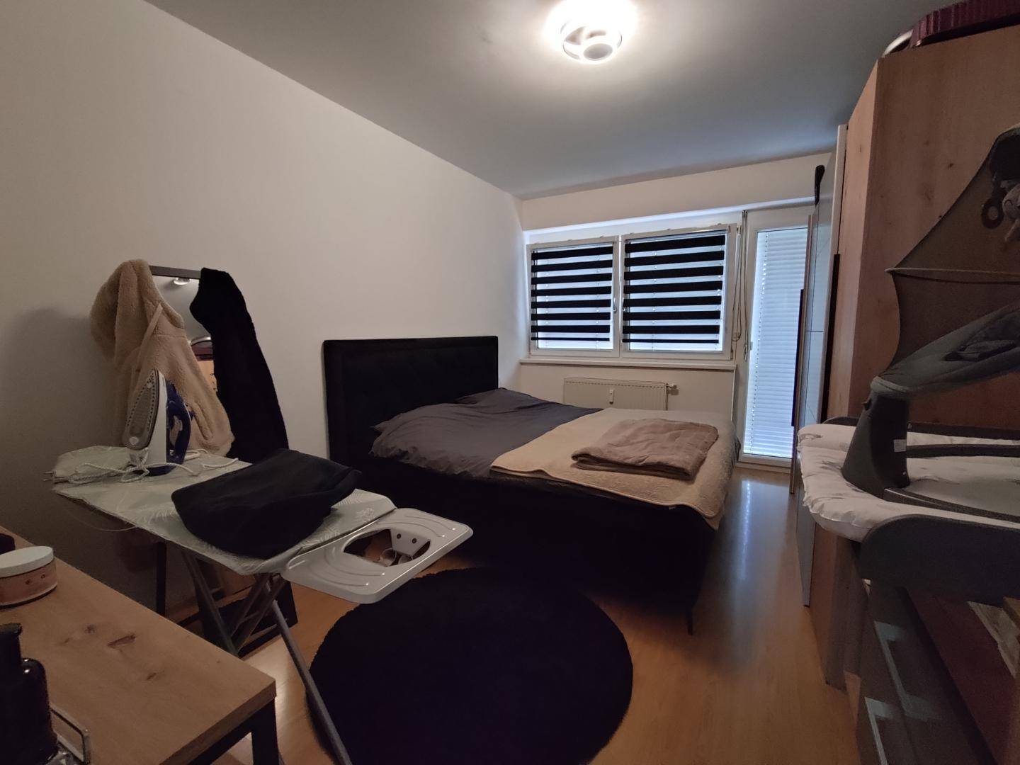 Schlafzimmer mit Doppelbett, Fenster und Holzmöbeln in schlichter Einrichtung.