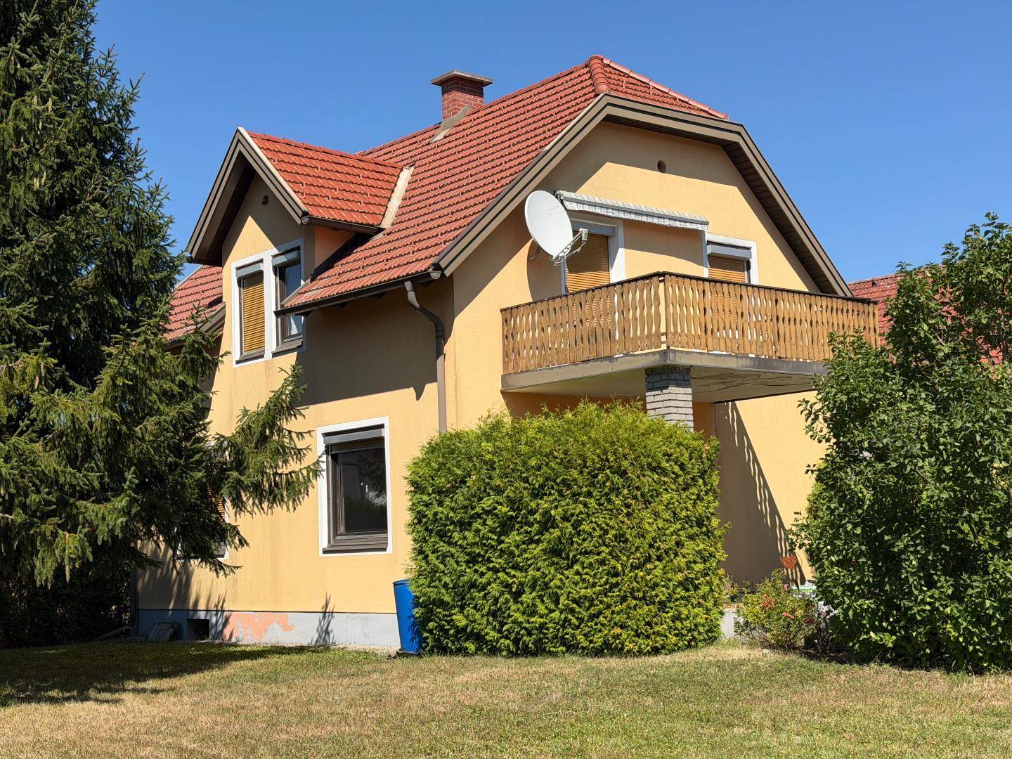 Seitenansicht eines Einfamilienhauses mit gelber Fassade, rotem Dach und einem Balkon im Obergeschoss.