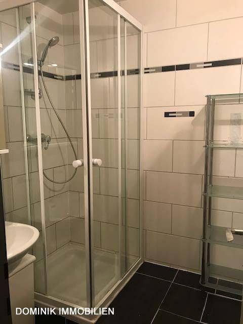 Badezimmer mit Glasduschkabine, weißen Wandfliesen und dunklem Boden, inklusive Regal.