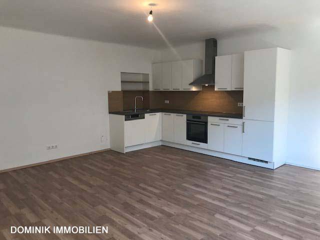 L-förmige Einbauküche mit weißen Schränken und Holzrückwand in einem offenen Wohnbereich mit Laminatboden.
