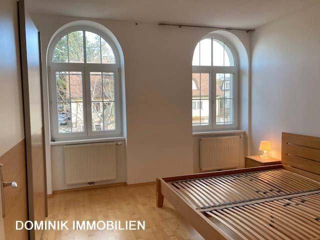 Schlafzimmer mit Holzbett, Parkettboden und zwei hohen Rundbogenfenstern.