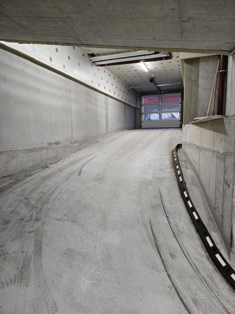 Gerade Rampe führt aus der Tiefgarage, umgeben von rohem Beton und technischen Leitungen.