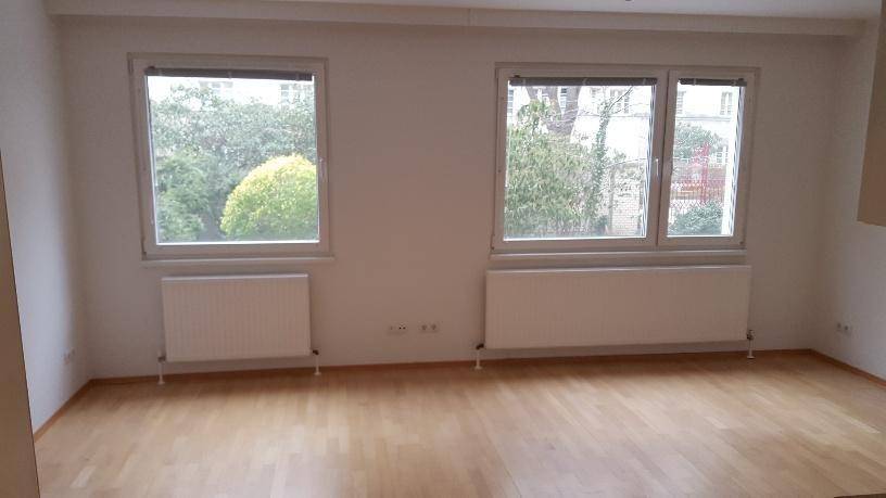 Leeres Wohnzimmer mit Parkettboden, zwei Fenstern und Heizkörpern unter den Fenstern.