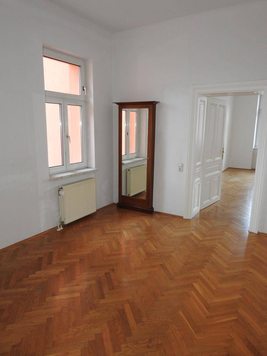 Helles Zimmer mit Fischgrätparkett, Fenster und einem eleganten Standspiegel.