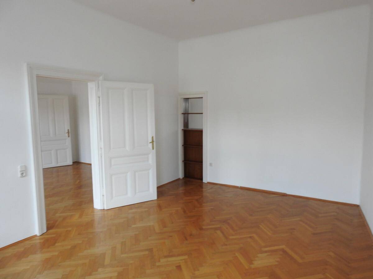Geräumiges Zimmer mit Fischgrätparkett, weiß gestrichenen Wänden und einer Nische mit Regal.