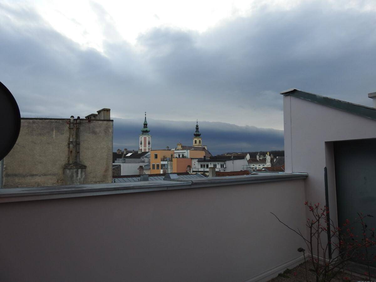Weitblick von der Terrasse über die städtische Bebauung mit zwei Kirchtürmen am Horizont unter dunklen Wolken.