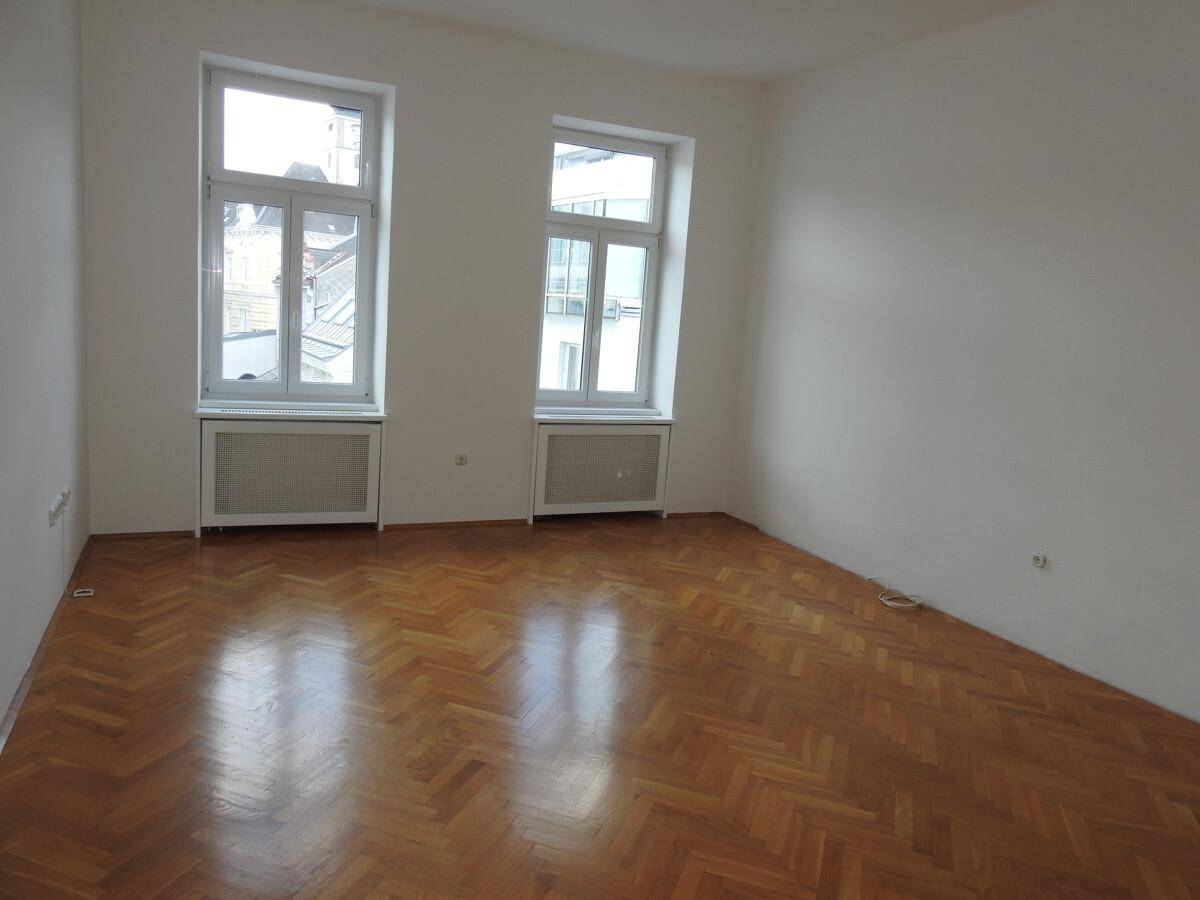 Geräumiges Zimmer mit zwei großen Fenstern, Fischgrätparkett und Heizkörpern unter den Fenstern.