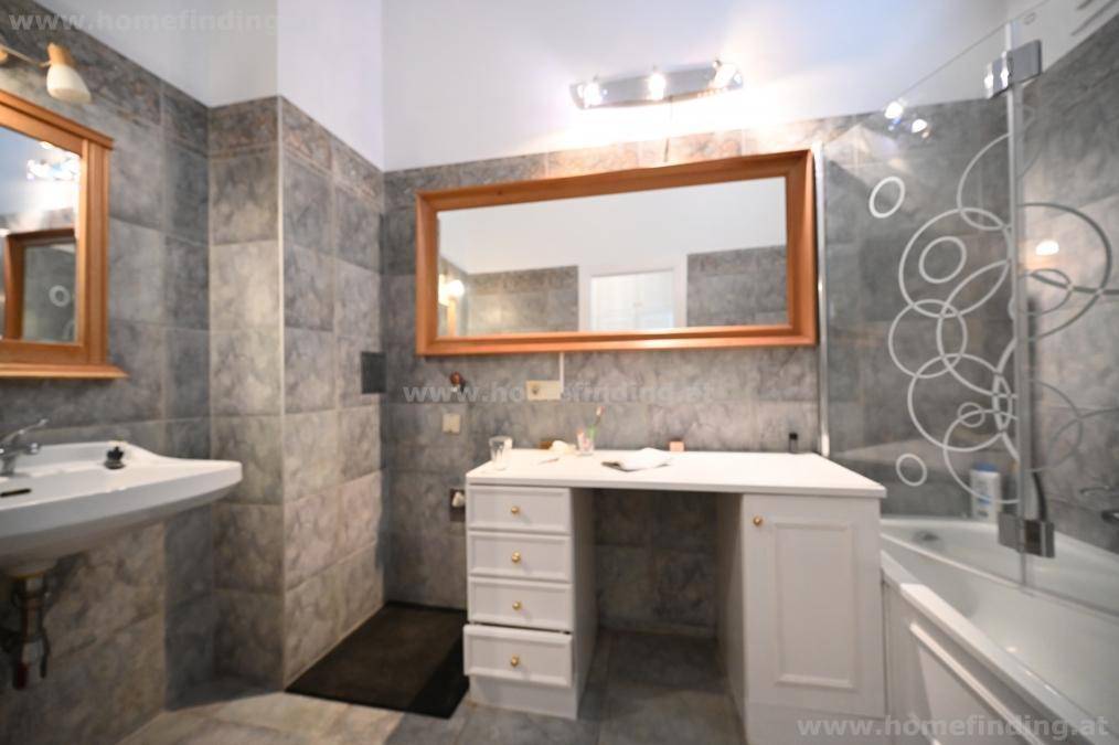 Badezimmer mit grauen Fliesen, Badewanne mit Duschwand und einem Waschtisch mit großem Spiegel.