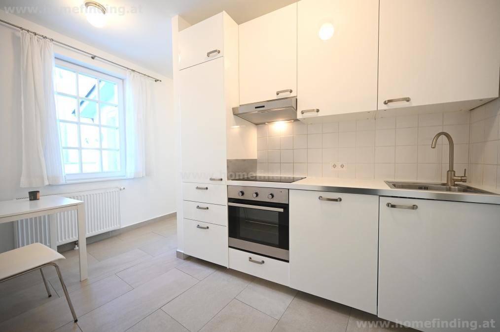Kleine, moderne Küche mit weißen Hochglanzschränken, integriertem Backofen und Fliesenboden.