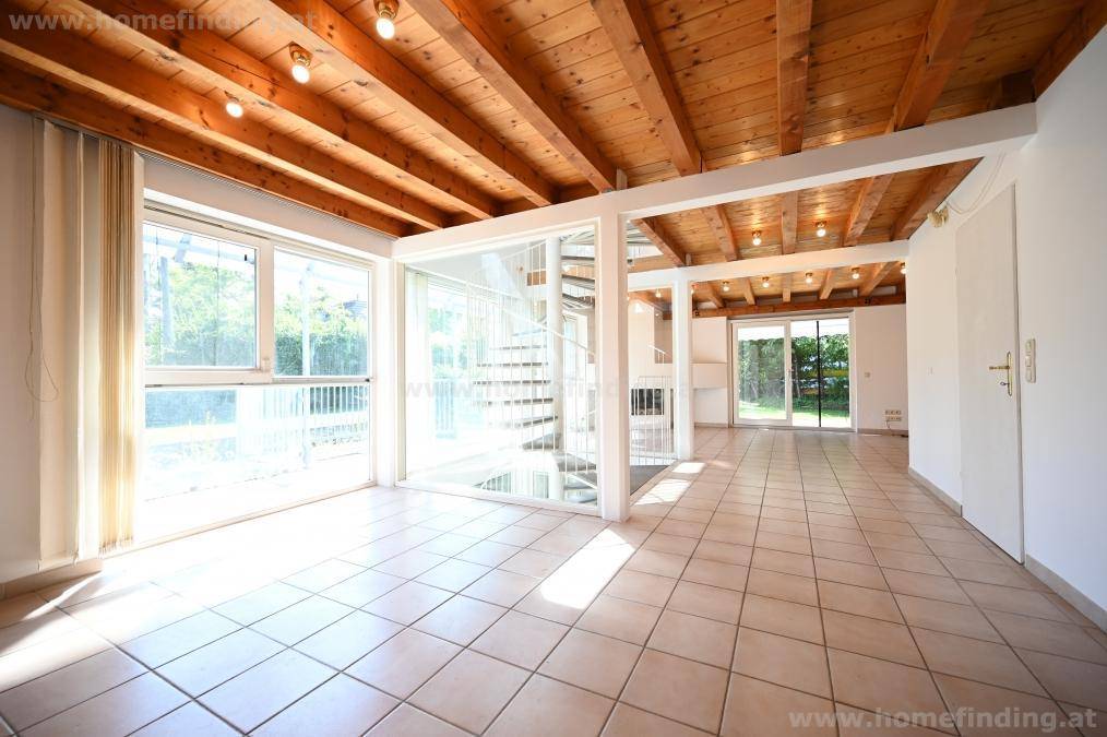 Heller Wohnbereich mit Fliesenboden, sichtbaren Holzdeckenbalken und einer eleganten Wendeltreppe aus Glas.