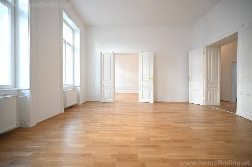 Großzügiges Zimmer mit hellem Parkettboden, zwei Fenstern und Zugang zu weiteren Räumen.