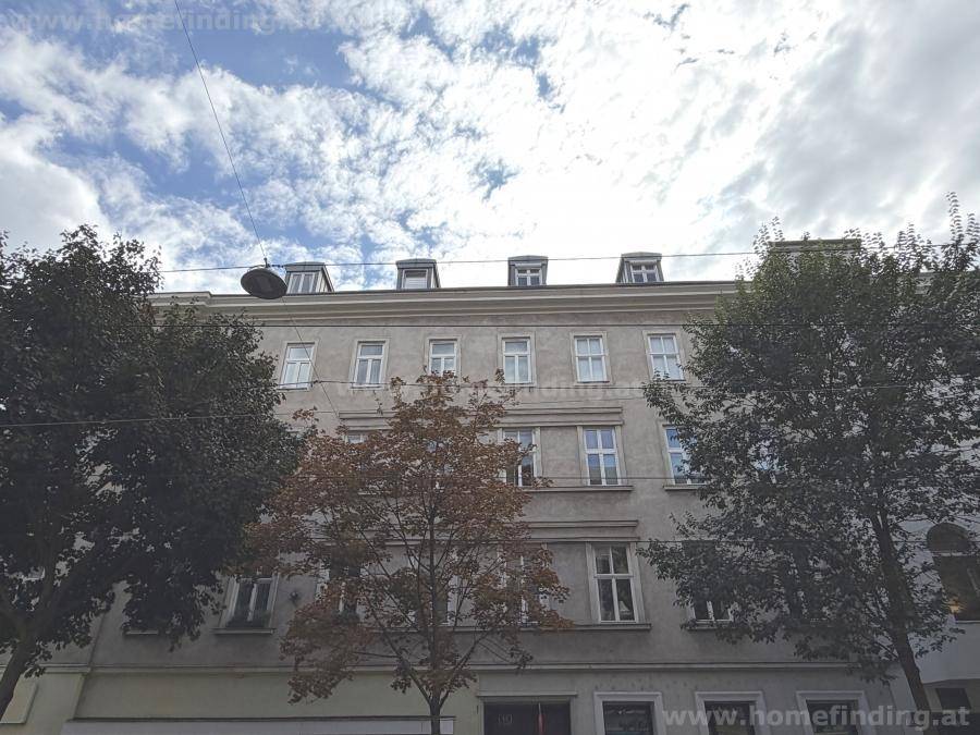 Klassische Fassade eines Altbaus mit vielen Fenstern und Bäumen im Vordergrund unter bewölktem Himmel.