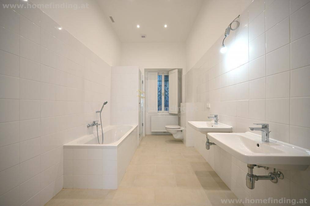 Geräumiges Badezimmer mit weißen Fliesen, zwei Waschbecken, Badewanne und Fenster für Tageslicht.