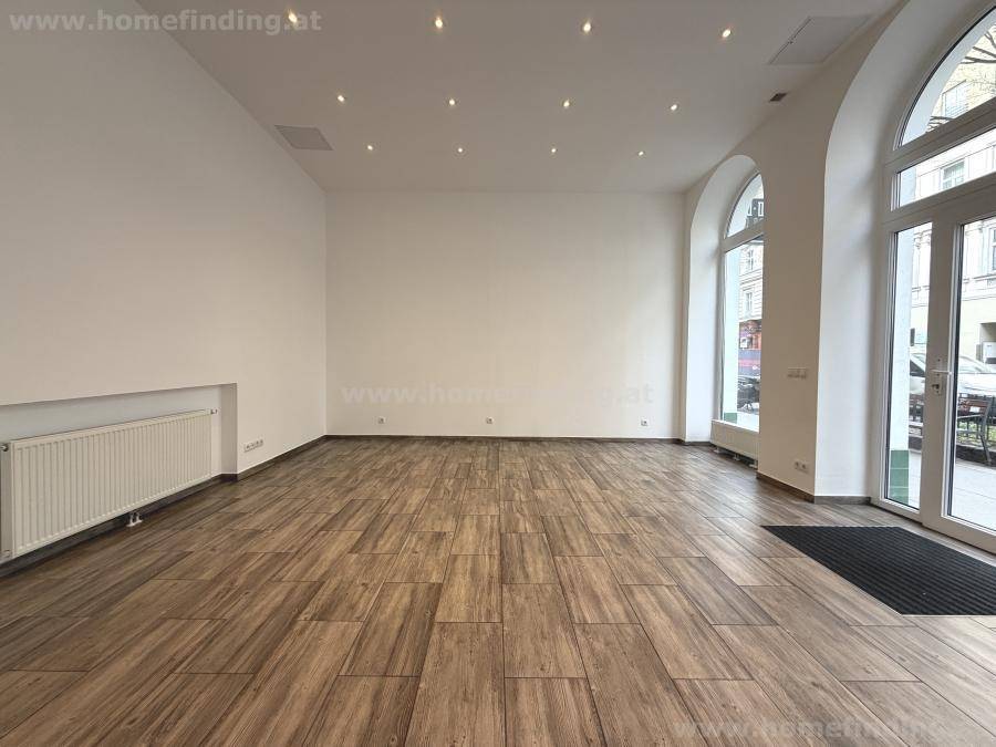 Lichtdurchfluteter Raum mit modernem Holzfliesenboden, weißen Wänden und Zugang zur Straße durch eine Glastür.