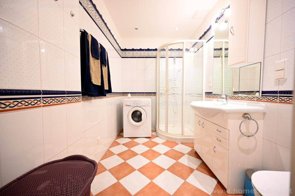 Badezimmer mit Eckdusche, Waschbeckenunterschrank, Waschmaschine und farbigen Fliesen.