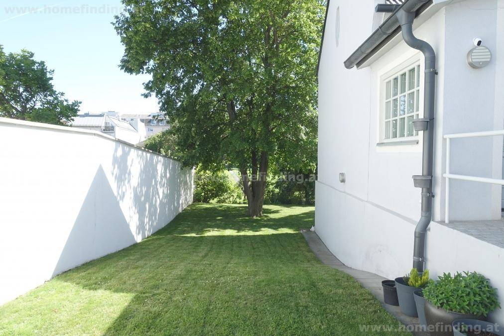 Gepflegte Rasenfläche entlang der Hausseite mit hoher weißer Mauer und einem großen Baum.