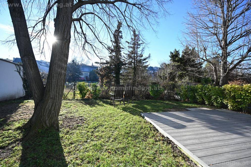 Sonniger Gartenbereich mit Rasenfläche, Bäumen und einer Holzterrasse zum Entspannen im Freien.