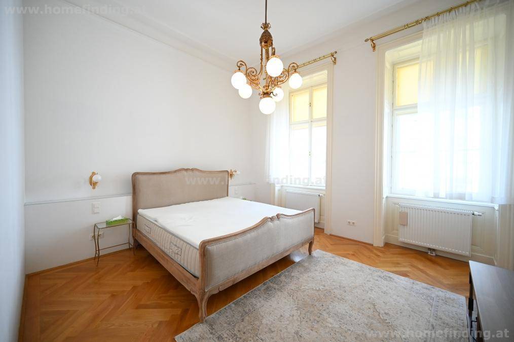 Klassisches Schlafzimmer mit elegantem Bett, Parkettboden und zwei großen Fenstern für viel natürliches Licht.