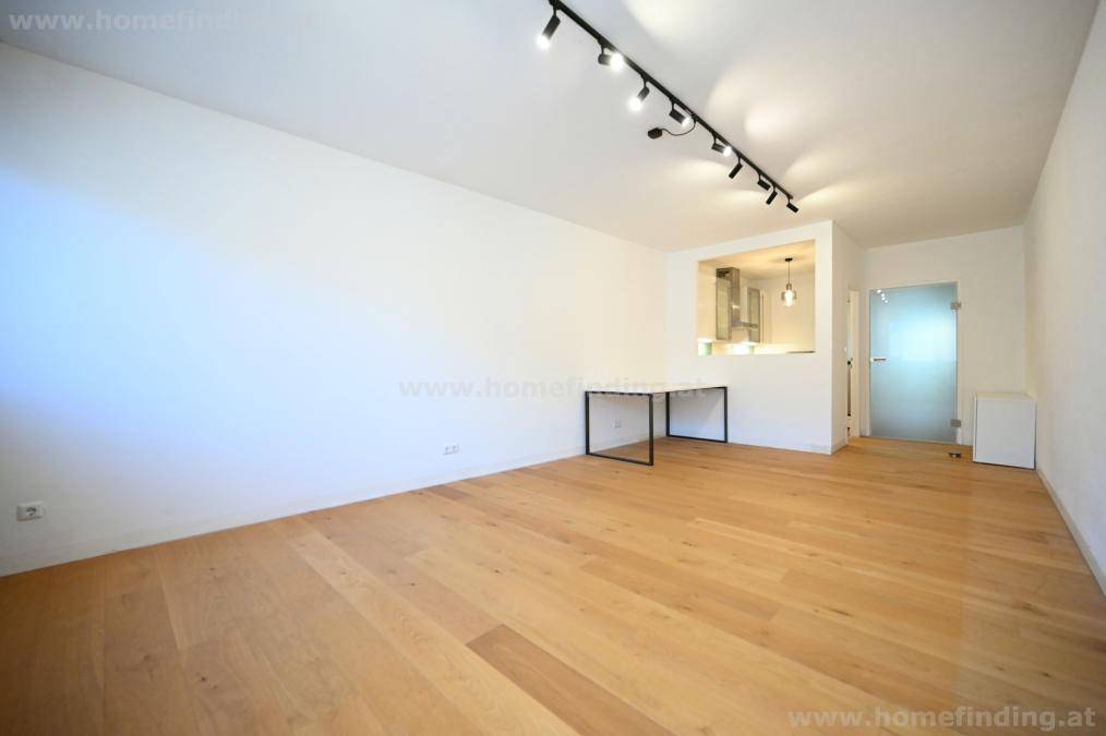 Heller Wohnbereich mit Holzboden, Blick zur Küche und einer modernen Glastür, die zu weiteren Räumen führt.