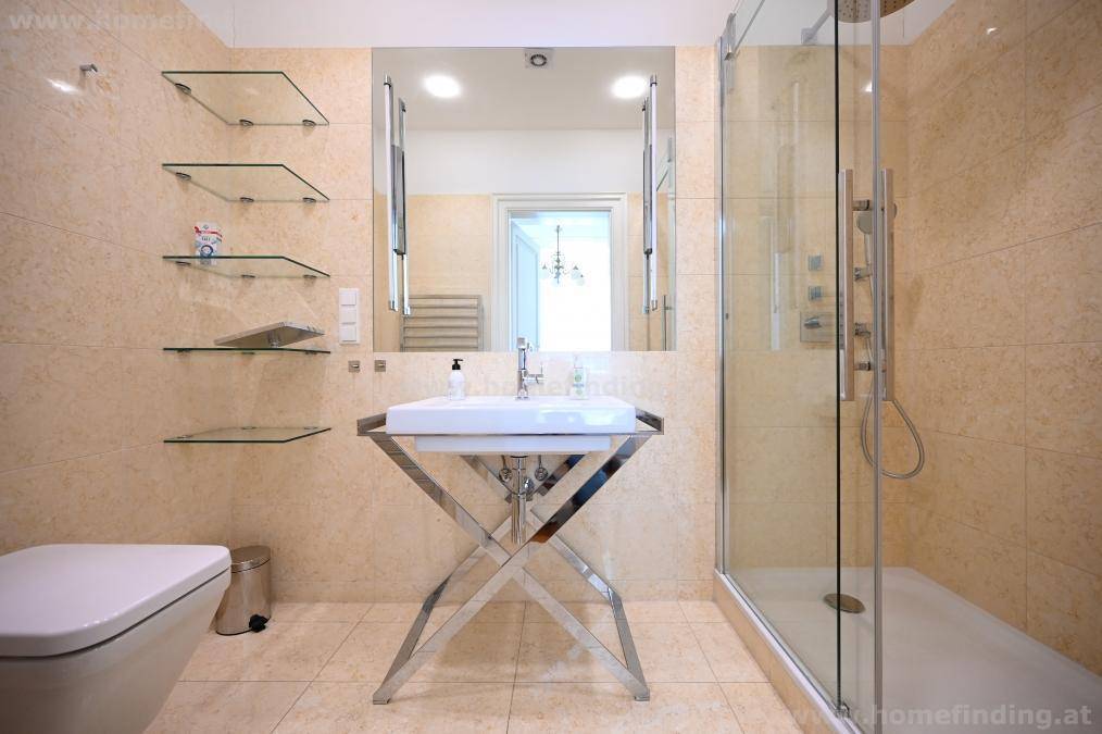 Helles Badezimmer mit beigen Fliesen, Duschkabine, Toilette und einem stilvollen Waschbecken auf Metallgestell.