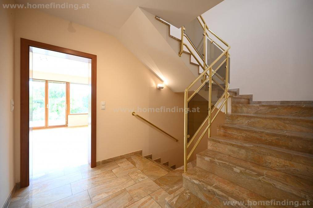 Heller Flur mit einer modernen Treppe, die Glas- und goldfarbene Geländer kombiniert, auf einem gefliesten Boden.
