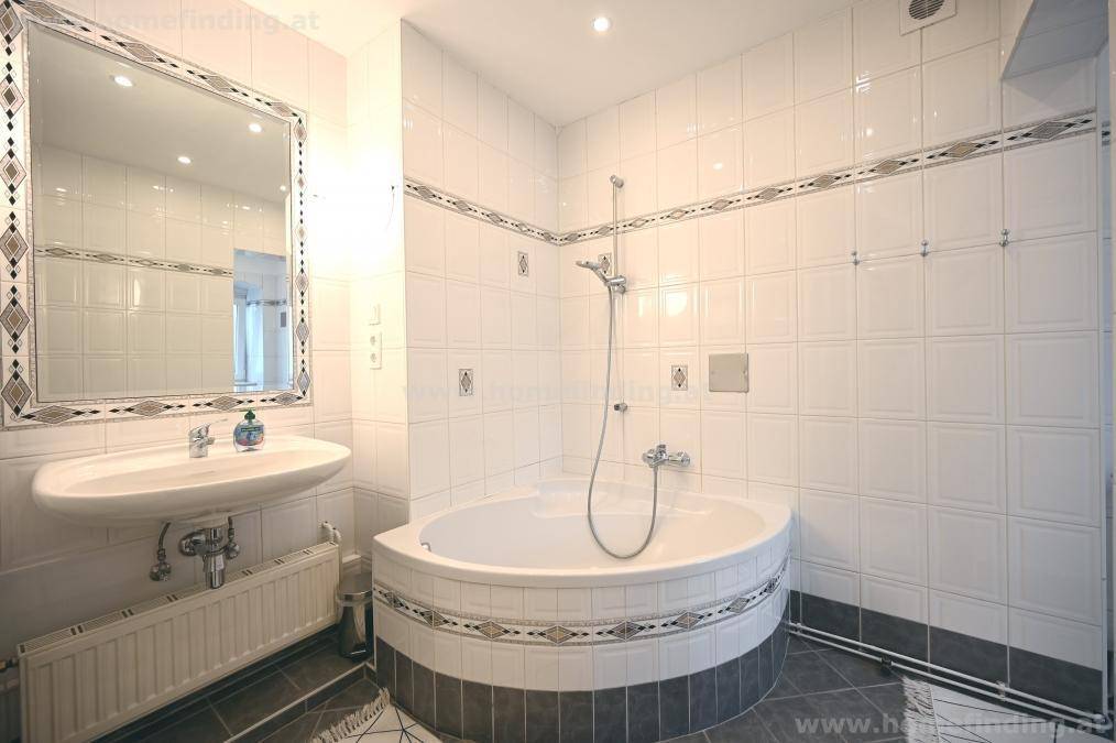 Badezimmer mit Eckbadewanne, Waschbecken und weißen Fliesen mit dekorativen Akzenten.