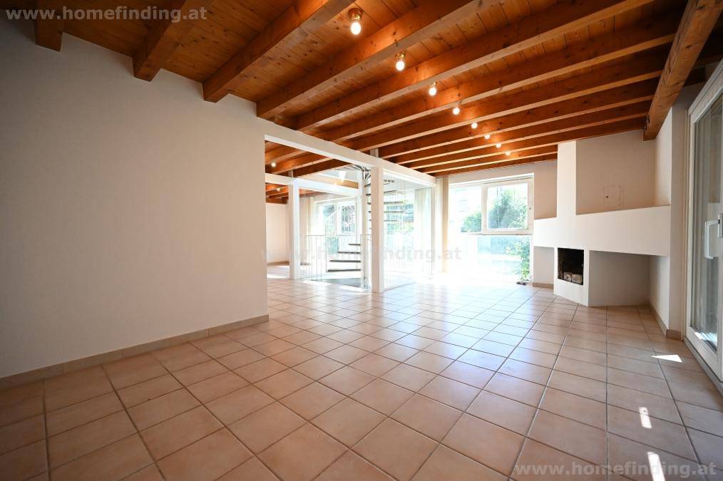 Geräumiger Wohnbereich mit Kamin, Fliesenboden, sichtbaren Holzdeckenbalken und viel Tageslicht.