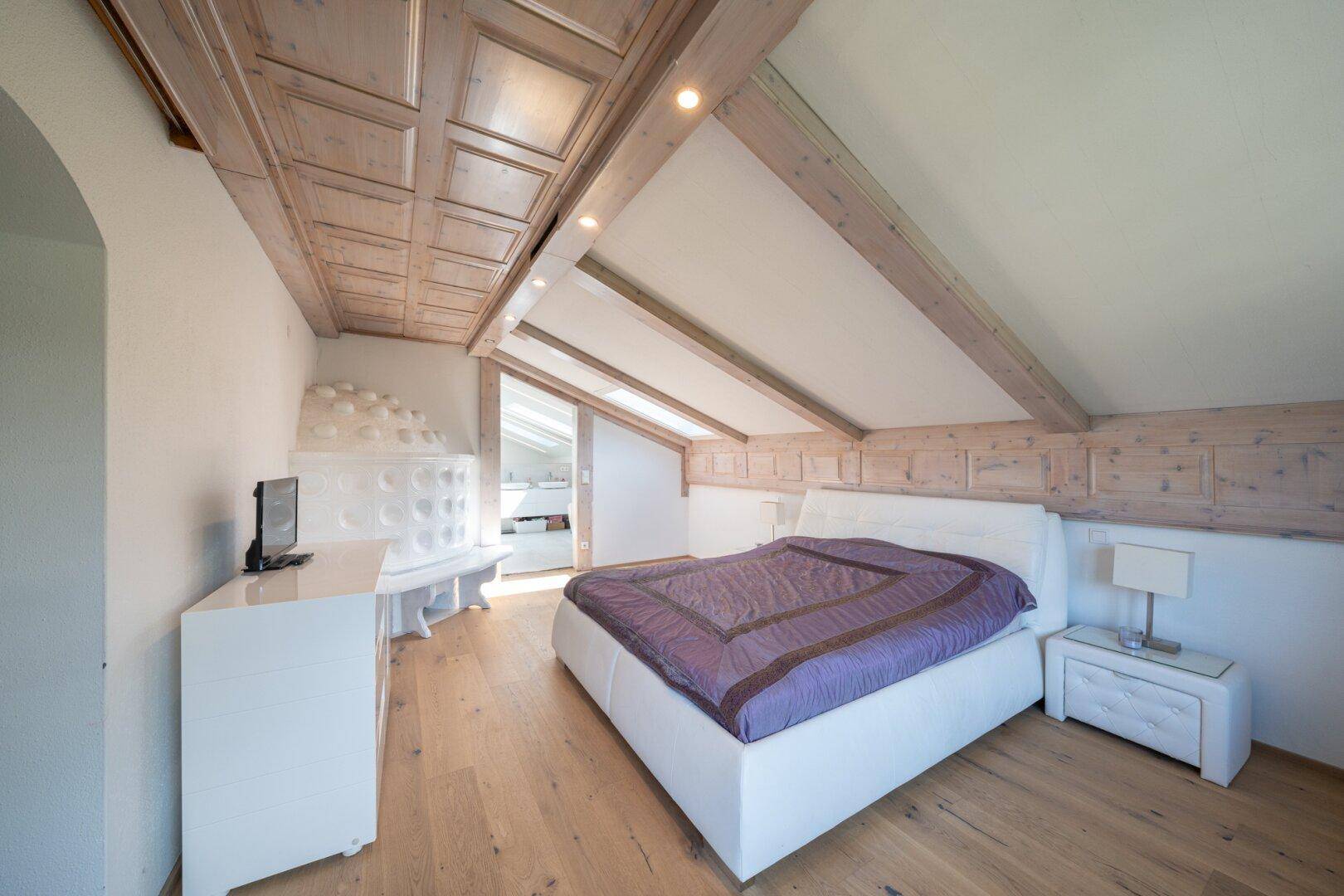 Helles Schlafzimmer im Dachgeschoss mit Holzbalkendecke, Kachelofen und Parkettboden.