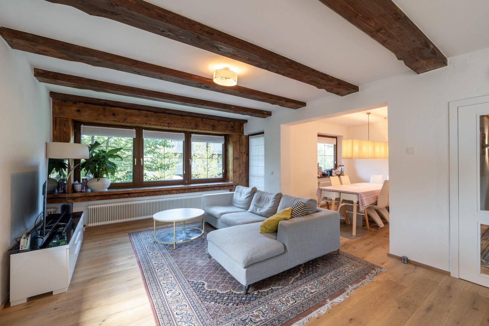 Gemütliches Wohnzimmer mit bequemer Couch, Holzbalken und großen Fenstern mit Blick ins Grüne.