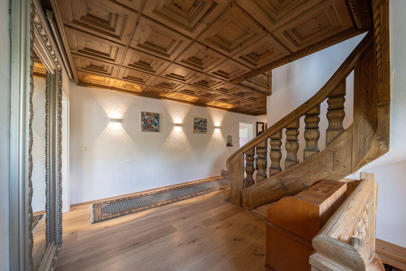 Breiter Flur mit kunstvoller Holztreppe, Kassettendecke und großem Spiegel.