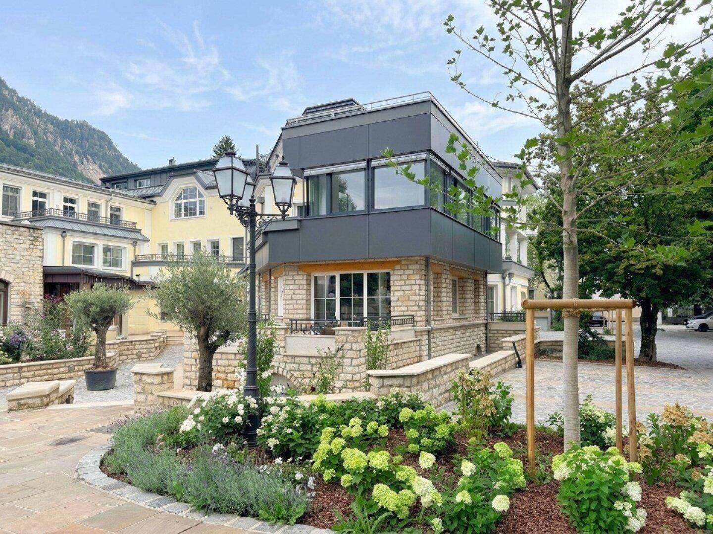 Modernes Gebäude mit Steinfassade und großen Fenstern, umgeben von einer gepflegten Gartenanlage und Bergen im Hintergrund.
