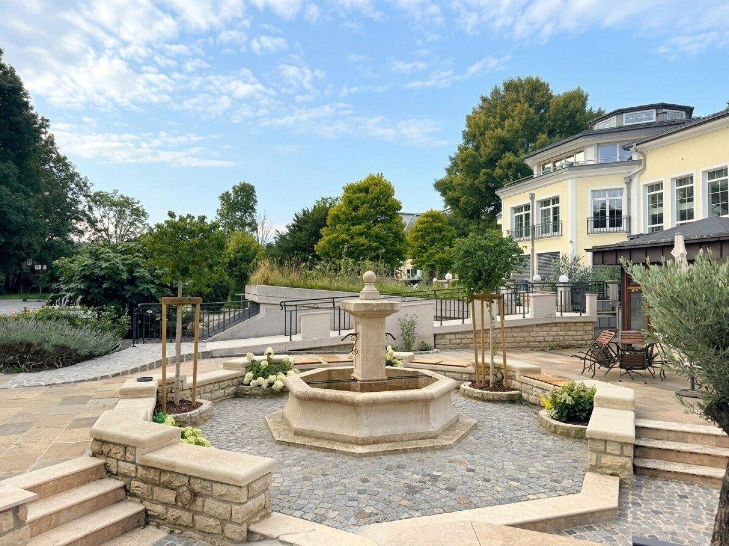 Attraktiver Innenhof mit einem zentralen Steinbrunnen, gepflasterten Wegen und üppiger Bepflanzung vor einem Gebäude.