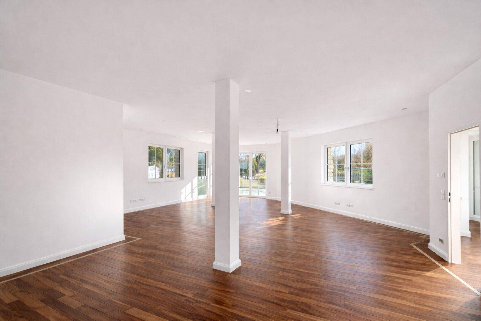 Heller, geräumiger Raum mit weißen Wänden, dunklem Holzfußboden und großen Fenstern, die viel Licht hereinlassen.