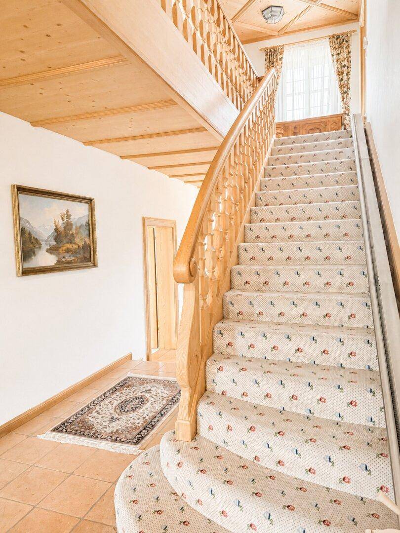 Treppenhaus mit gemustertem Teppich, Holzgeländer und Holzbalkendecke, führt in die obere Etage.