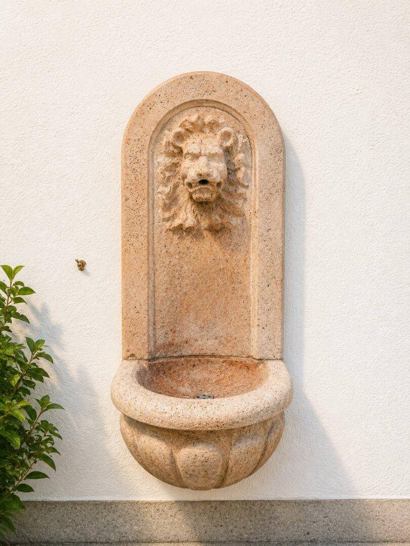 Dekorative Steinwandfontäne mit Löwenkopfmotiv an der weißen Hausfassade.