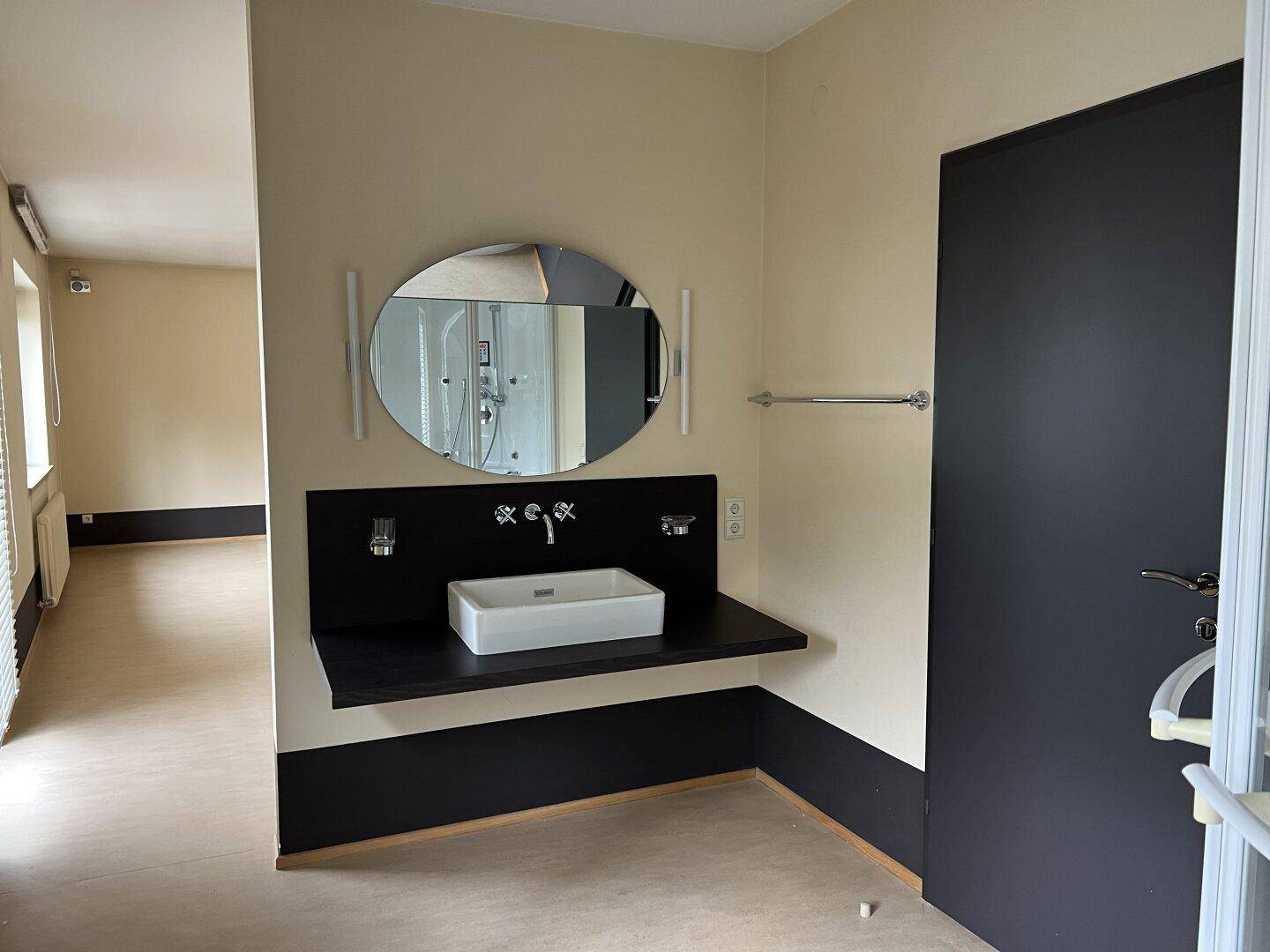 Badezimmerbereich mit modernem Waschbecken, ovalem Spiegel und indirekter Beleuchtung, Duschkabine im Spiegel sichtbar.