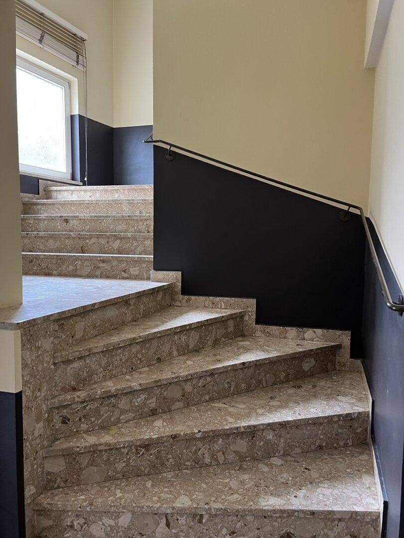 Treppenhaus mit Terrazzo-Stufen und schlichtem Metallgeländer, führt zu den oberen Etagen.