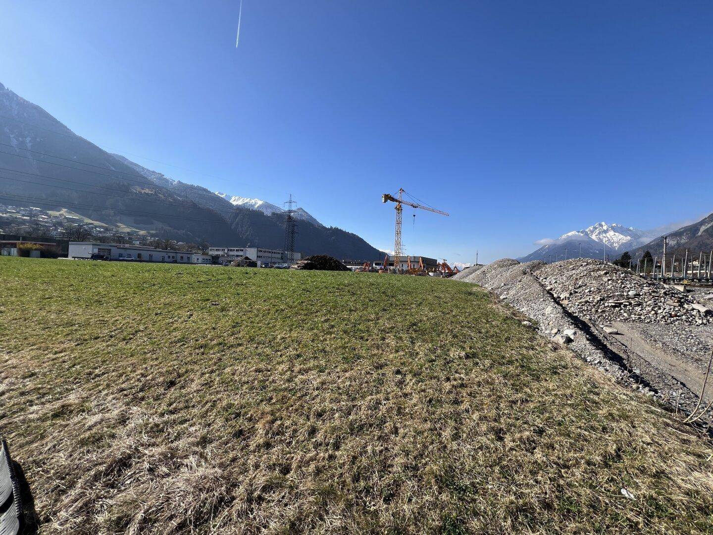 Blick über ein weites Feld auf schneebedeckte Berge und eine Baustelle mit Kran in der Ferne.