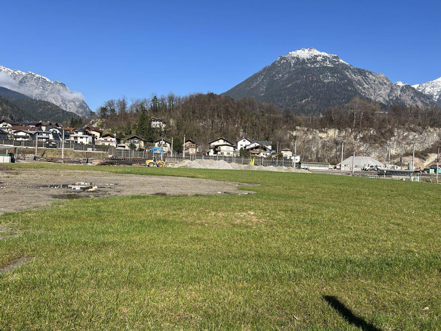 Weitläufiges grünes Feld mit einer Reihe von Häusern und schneebedeckten Bergen im Hintergrund.
