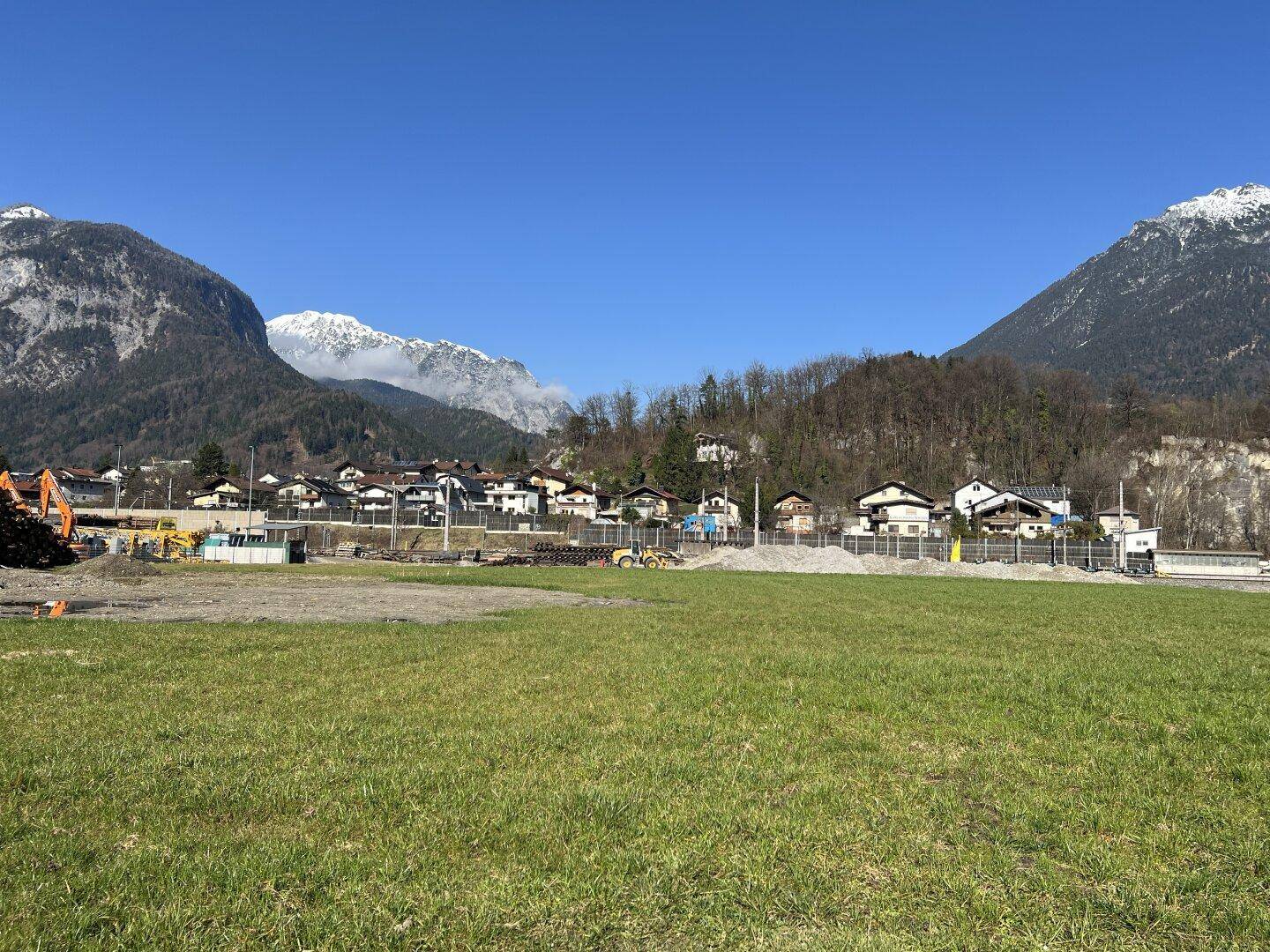 Blick über ein grünes Feld auf eine Siedlung mit Häusern und majestätischen schneebedeckten Bergen.