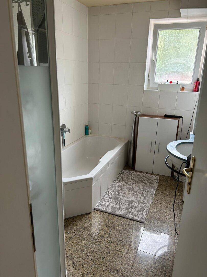Badezimmer mit Badewanne, Waschbecken und Fenster, ausgestattet mit Fliesenboden in Granitoptik.