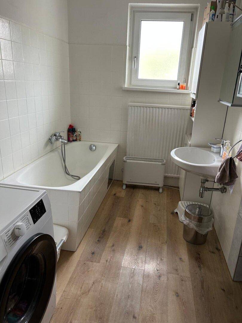 Badezimmer mit Badewanne, Waschmaschine und Fenster, hell gefliest und funktional eingerichtet.