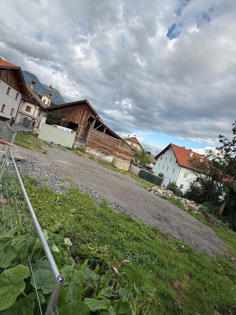 Schotterfläche eines Grundstücks mit angrenzenden Gebäuden und Bergpanorama unter bewölktem Himmel.